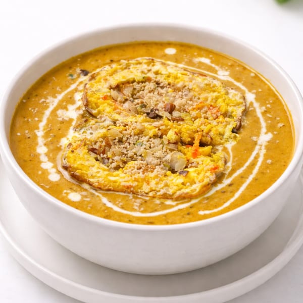 Close-up of a bowl of creamy orange soup with a yellow omelet and nuts on top.