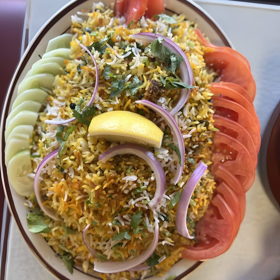 A vibrant Indian biryani dish, garnished with sliced cucumbers, tomatoes, red onions, and a lemon wedge.