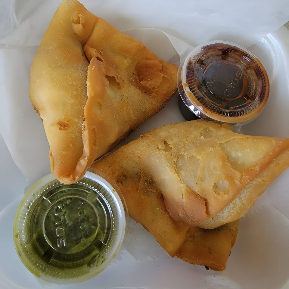 Two golden-brown samosas with green and brown dipping sauces in plastic containers.