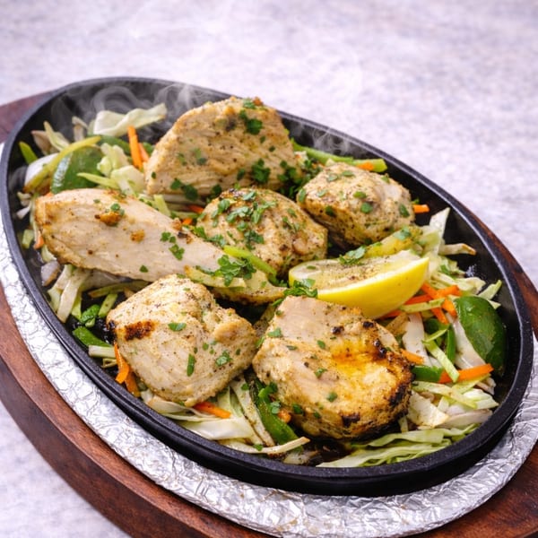 Steaming chicken tikka pieces with vegetables and a lemon wedge in a sizzling cast iron platter.